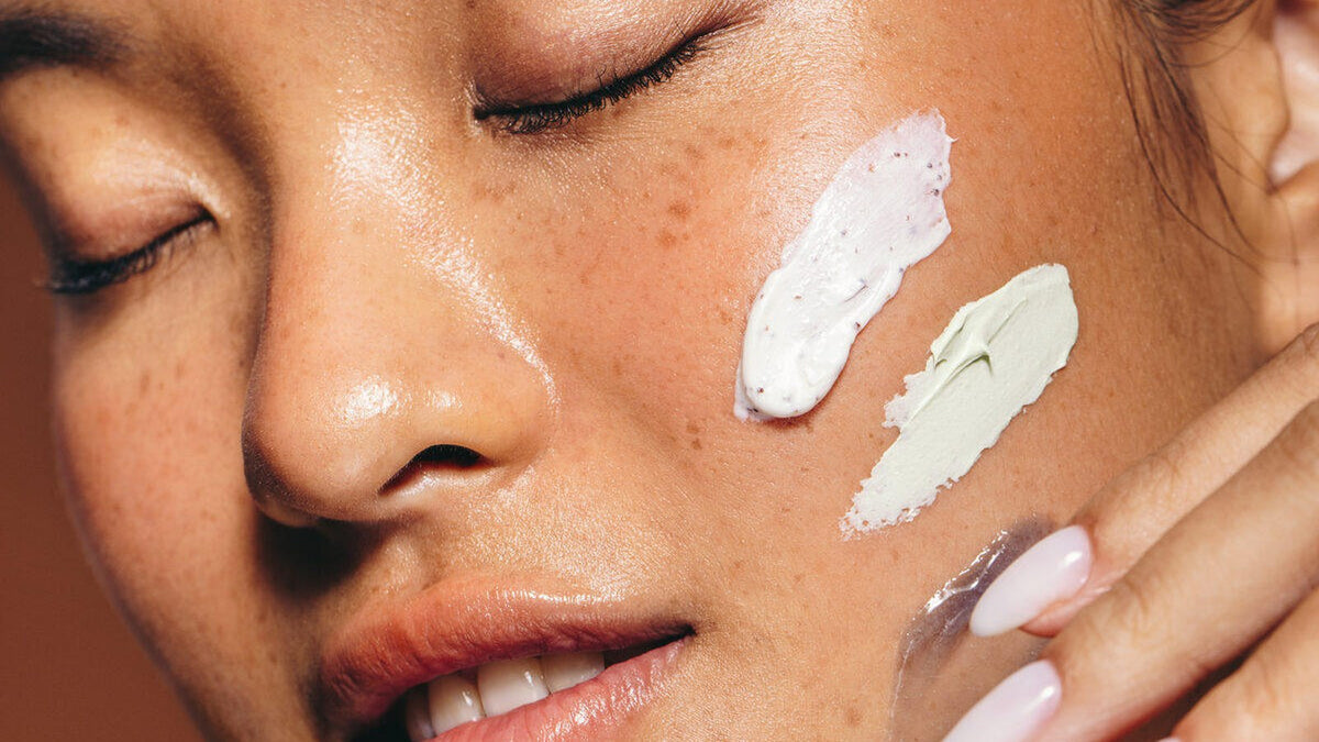 A woman with her eyes closed and three swatches of skincare on her cheek, one white cream, one sage green and one clear gel